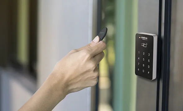 Mão aproximando um cartão de um controlador de acesso Intelbras na porta de vidro de uma empresa